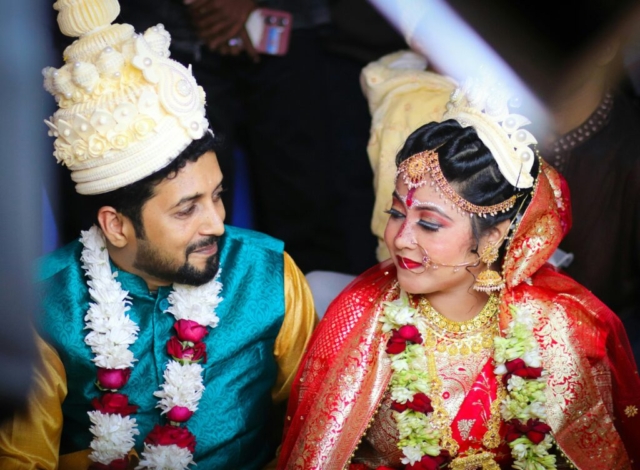 A joyful Indian couple in traditional wedding clothing adorned with jewelry and flowers.