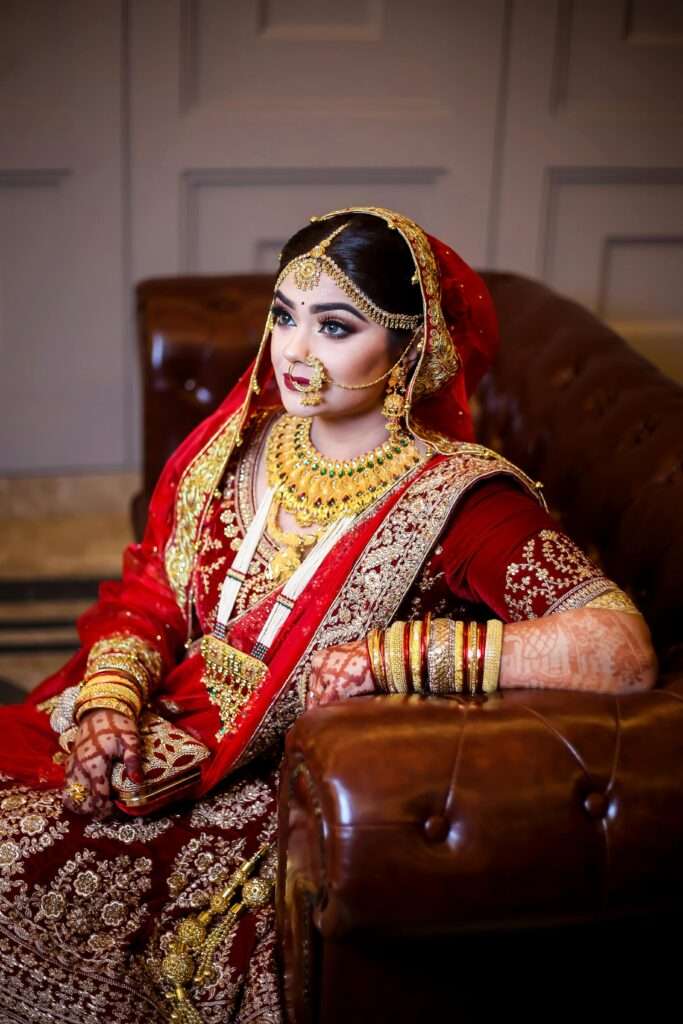 South Asian bride in traditional red and gold attire with henna and jewelry indoors.